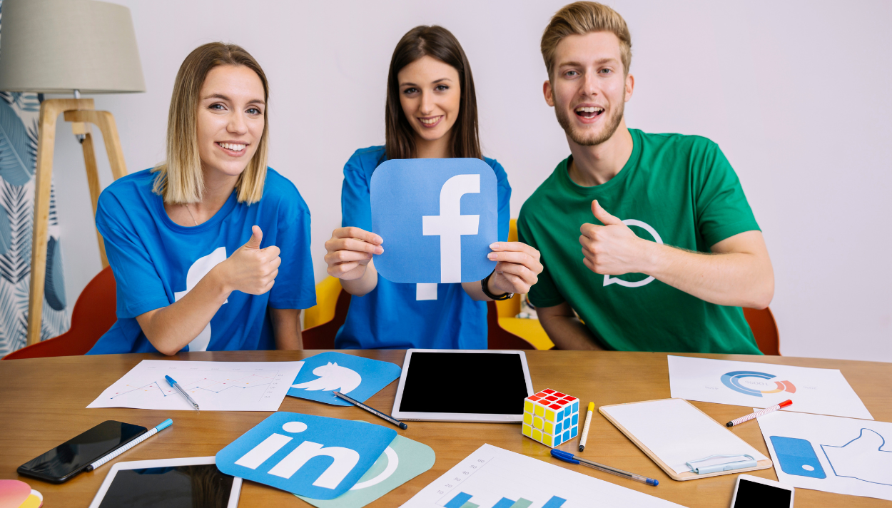 Three youngsters are posing for a camera with thumbs up. The girl in the middle is holding a Facebook logo made on paper.